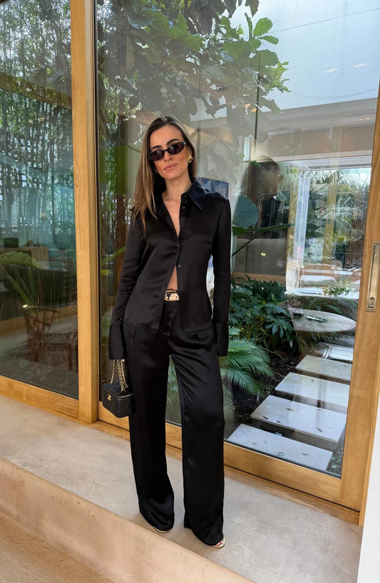 A woman wears the Tara Satin Long Sleeve Shirt Black paired with wide-leg trousers, a snakeskin belt, gold earrings, and a black quilted Chanel bag.