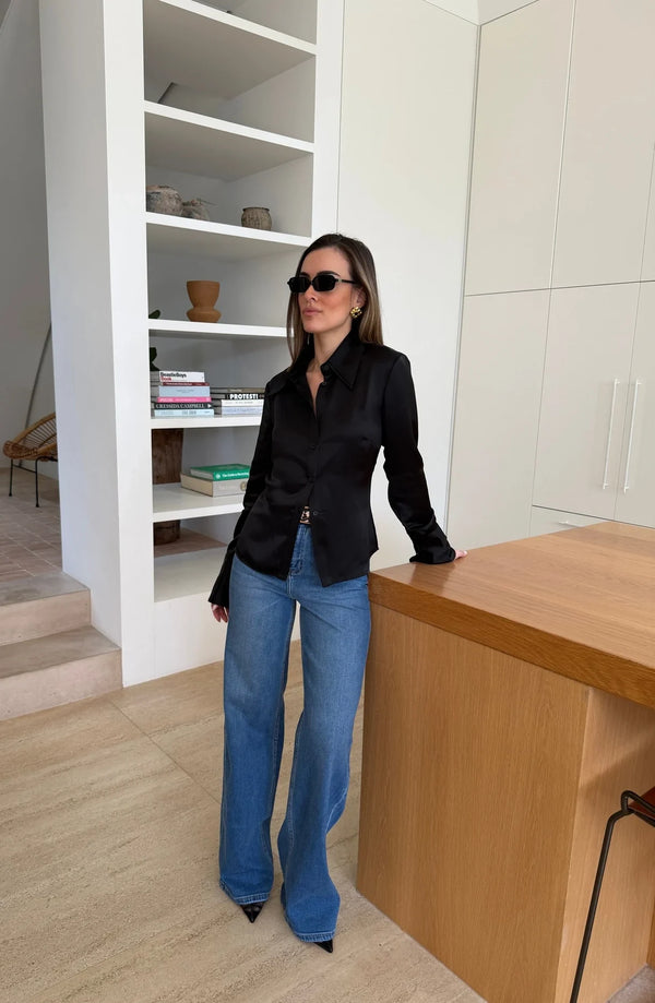 A woman wearing a black Tara Satin Long Sleeve Shirt and wide-leg jeans leans against a wooden kitchen island in a modern, minimalist home.
