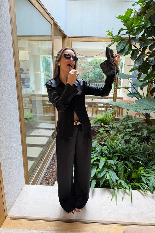 A woman in a Tara Satin Long Sleeve Shirt Black applies lipstick using her quilted black handbag in a bright, modern atrium.