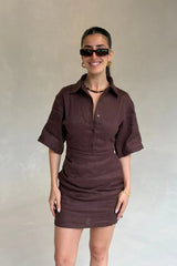 Woman wearing a dark brown button-down Vida Mini Dress with short sleeves and gold accessories against a neutral wall.
