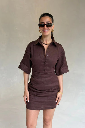 Woman wearing a dark brown button-down Vida Mini Dress with short sleeves and gold accessories against a neutral wall.