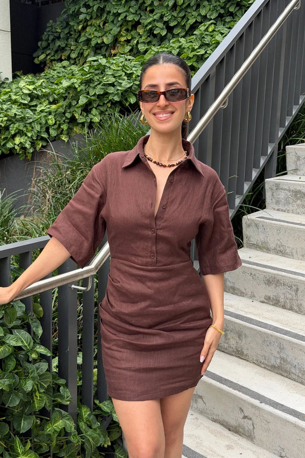 A young woman confidently poses on concrete stairs wearing a chocolate brown Vida Mini Dress with tortoise-shell sunglasses and gold hoop earrings.