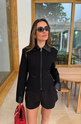 A woman wears the Winnie Fitted Zip Jacket Black with matching shorts, gold hoop earrings, dark sunglasses, and a red handbag in a bright, modern room.