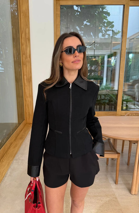 A woman wears the Winnie Fitted Zip Jacket Black with matching shorts, gold hoop earrings, dark sunglasses, and a red handbag in a bright, modern room.