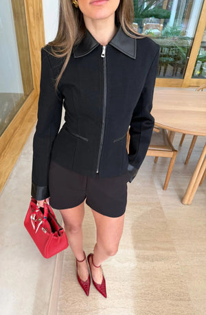 A woman wears the Winnie Fitted Zip Jacket Black with black shorts, red handbag, and matching red crocodile-embossed heels in a modern interior.