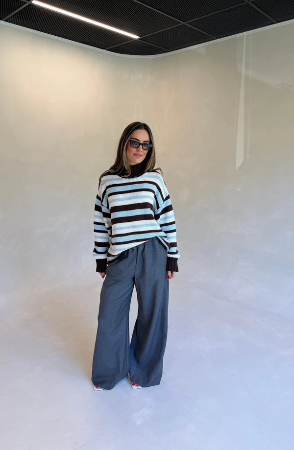 A woman wears Zoe Relaxed Suit Pant Grey with a striped high-neck sweater and dark sunglasses against a minimalist off-white backdrop.
