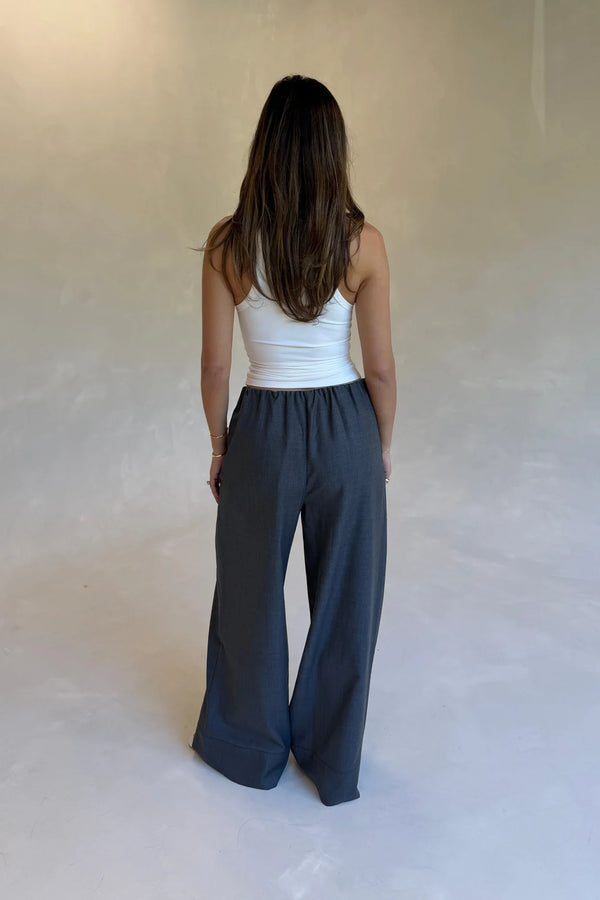 A woman stands facing away wearing a white halter tank and Zoe Relaxed Suit Pant Grey wide-leg trousers in a minimalist studio.