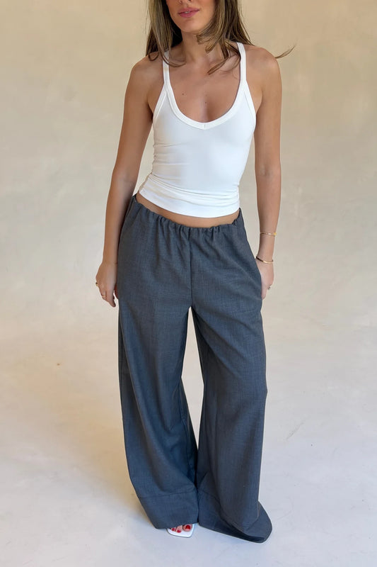 A woman wears relaxed wide-leg grey Zoe suit pants with a white tank top and white wedge sandals in a minimalist studio.