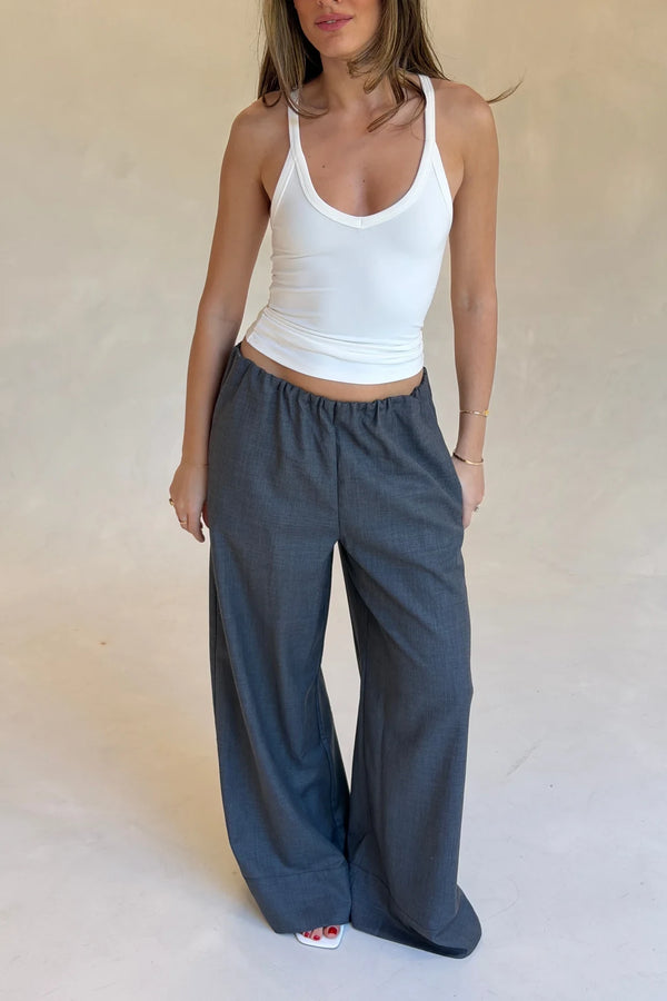 A woman wears relaxed wide-leg grey Zoe suit pants with a white tank top and white wedge sandals in a minimalist studio.